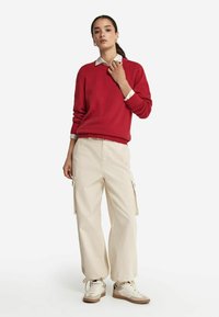 Roter Strickpullover mit Rundhalsausschnitt, beige Cargo-Hosen mit Seitentaschen und weiße Sneakers mit braunen Akzenten. Einfaches und lässiges Outfit.