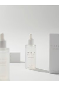 Bottiglia contagocce in vetro trasparente contenente siero, con tappo contagocce bianco, etichettata "NEEDLY Ampoule", circondata da un packaging minimal.