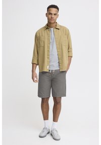 Tenue décontractée comprenant une chemise beige à boutons, un t-shirt gris, un short gris et des baskets grises avec des chaussettes blanches. Design simple et textures douces.
