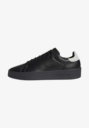 Zapatilla deportiva de cuero negro con una textura lisa, que presenta una suela negra gruesa y perforaciones sutiles, acentuada por una pestaña blanca en el talón.
