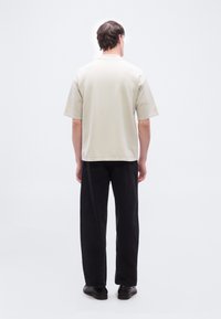 Άνδρας που φοράει μπεζ πουκάμισο με κοντά μανίκια, μαύρο παντελόνι και μαύρα παπούτσια, στέκεται με την πλάτη προς το μέρος μας μπροστά σε ένα απλό ανοιχτό φόντο.