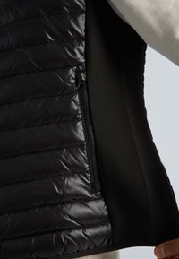 Gilet noir isolé avec des panneaux matelassés, un intérieur lisse et un détail de fermeture éclair sur le côté, contrastant avec la doublure en tissu doux.