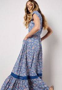 Robe maxi florale bleue avec des motifs roses et blancs, manches courtes, taille froncée et bord en dentelle au bas. Tissu fluide avec des poches.