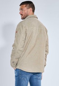 Hellbeige Cordjacke aus Cord mit Standardkragen, langen Ärmeln und einem Aufnäher am linken Arm. Kombiniert mit blauen Jeans aus Denim.