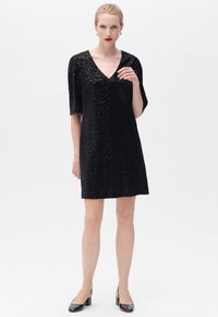 Vestido negro de lentejuelas con escote en V, mangas cortas y un corte suelto, combinado con tacones negros. Textura suave con detalles brillantes.