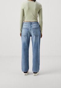 Svetle zelený dlouhý rukáv tričko v kombinaci s volnými světle modrými džínami. Džínsy mají dvě zadní kapsy a rovný střih. Viditelné bílé tenisky.