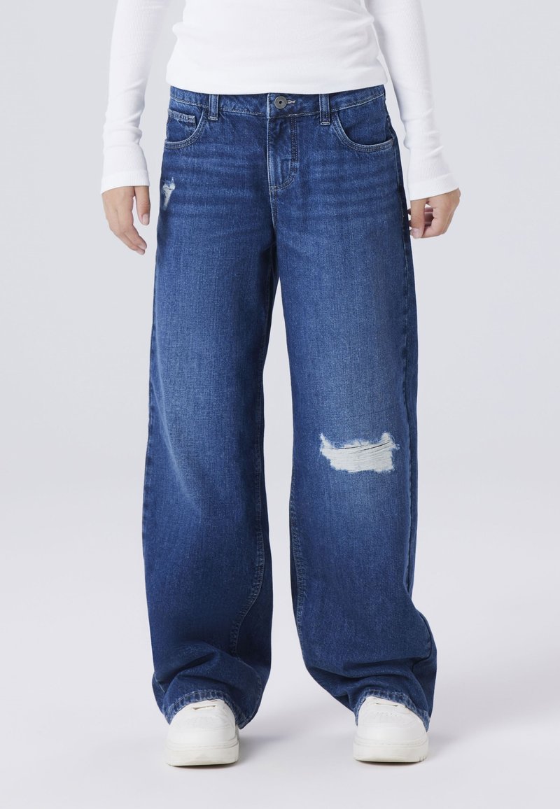 Jeans de mezclilla azul de pierna ancha con un acabado desgastado, que presentan un parche deshilachado en la rodilla izquierda y un estilo tradicional de cinco bolsillos.