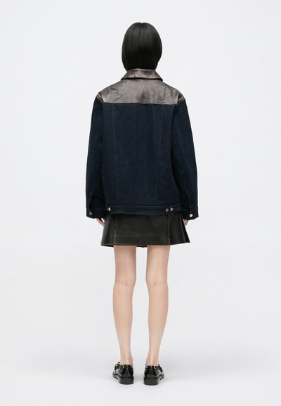 Σκούρο τζάκετ από τζιν με καφέ δερμάτινες τιράντες στους ώμους, με άνετη εφαρμογή και κουμπώματα με κουμπί στο τελείωμα, συνδυασμένο με δερμάτινη φούστα.