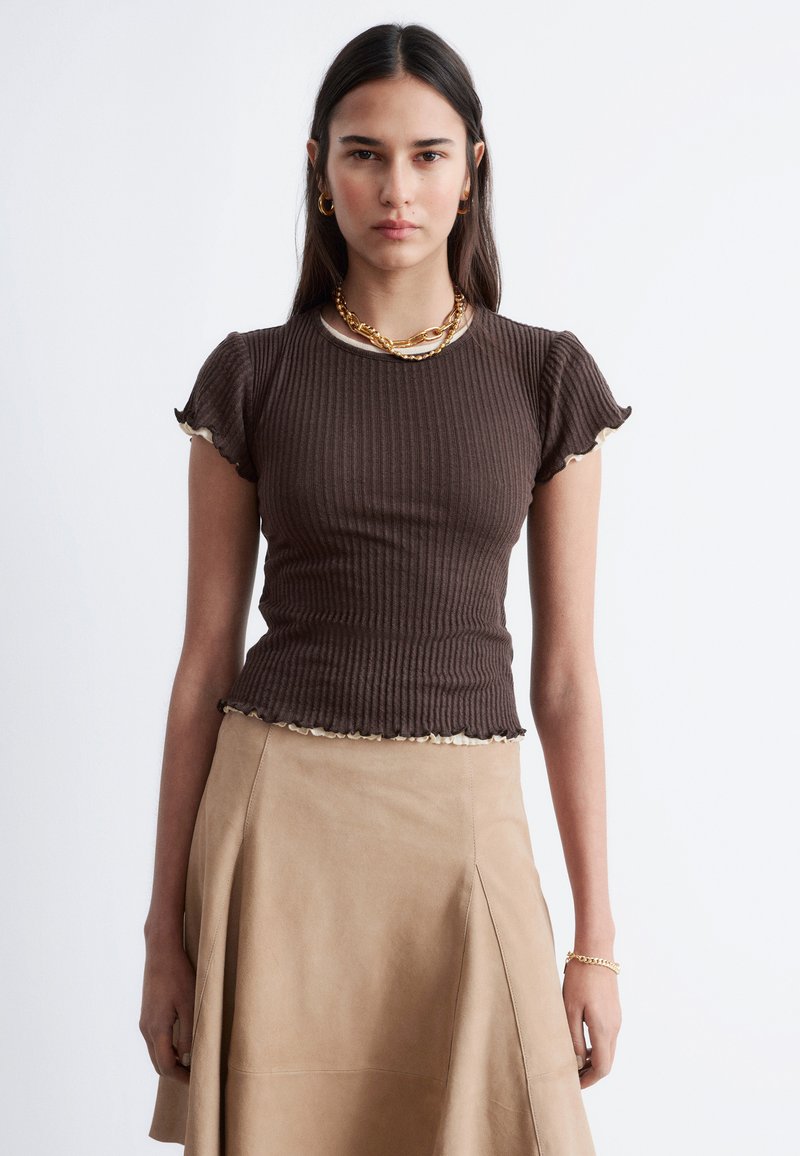 Kvinna som bär en figursydd brun ribbad topp med kort ärm och krämfärgad kant, beige A-linjig kjol, guldkedjehalsband, stora ringformade örhängen och armband.