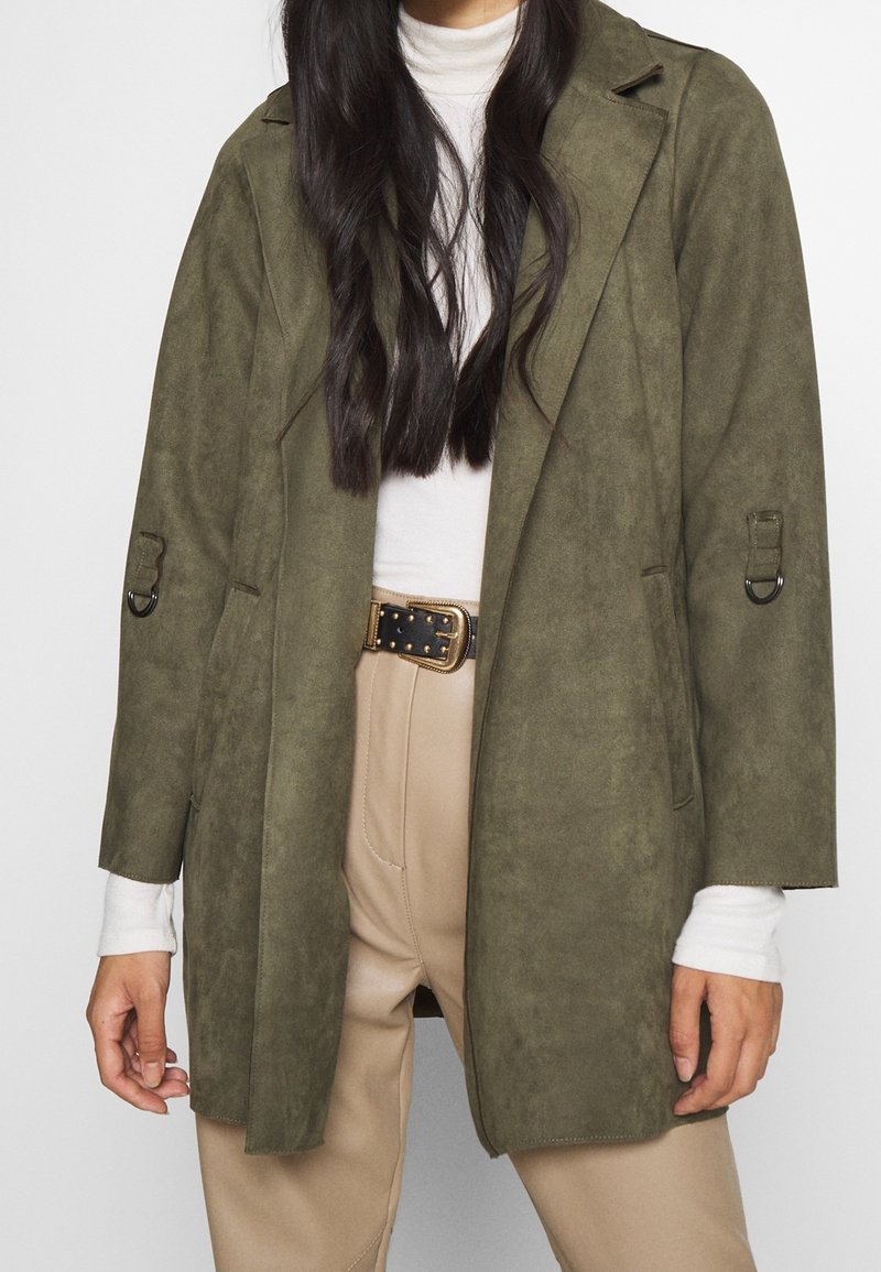 Manteau en daim vert olive avec une coupe décontractée, présentant un col cranté, des manches longues et des passants de ceinture. Porter par-dessus un turtleneck léger et un pantalon beige.