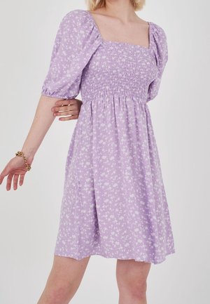 Vaaleatukkainen nainen, joka pukeutunut lilaiseen kukkakuosiseen mekkoon, jossa on puffihihat ja röyhelöity yläosa, poseeraa hieman ojennetuin käsin.