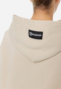 Felpa beige con un grande cappuccio morbido. Presenta un'etichetta nera PEGADOR cucita sul collo della schiena. Il tessuto appare liscio e strutturato.