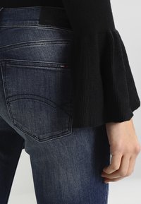 Jeans azul oscuro con corte ajustado, que presentan una textura sutilmente desgastada, bolsillos traseros con costura curva y una etiqueta negra en la cintura.