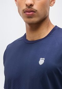 Νεαρός άνδρας με ελαφρύ μουστάκι, φοράει μπλε σκούρο αθλητικό πουκάμισο K-Swiss με λογότυπο στο στήθος, εμφανίζεται από τους ώμους μέχρι τα χείλη.