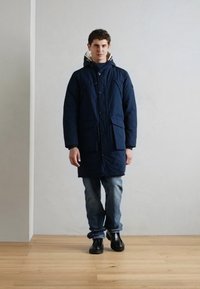 Parka acolchada de color azul marino con capucha, grandes bolsillos frontales y cierre de botones, combinada con jeans azules y zapatos negros sobre un suelo de madera.