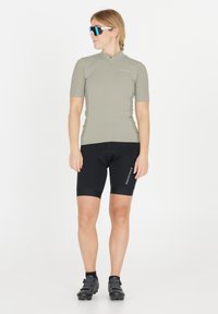 Nainen, jolla on beige lyhythihainen pyöräilypaita, mustat pyöräilyshortsit, mustat pyöräilykengät ja heijastavat urheilulasit, seisoo valkoisella taustalla.