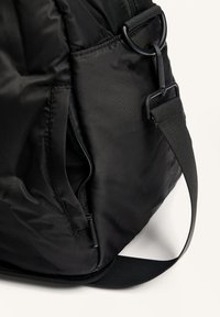 Sac en tissu noir avec une poche zippée partiellement ouverte et une sangle réglable attachée à un anneau métallique.