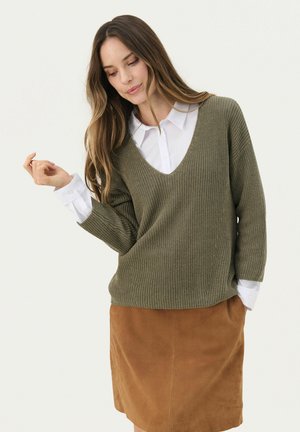 Olive green knitted sweater with a deep V-neck, featuring loose sleeves, worn over a white button-up shirt, paired with a brown suede skirt.