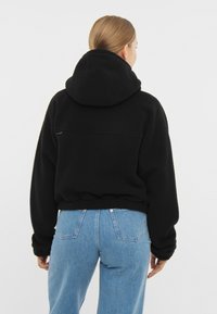 Felpa nera con cappuccio e collo alto, con una vestibilità comoda e polsini a coste. Dettaglio della tasca posteriore e texture morbida. Abbinata a jeans blu.