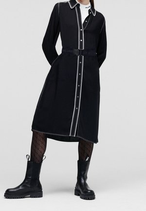 Person trägt ein schwarzes, langärmeliges, knöpfbares Kleid mit weißen Nähten, gemusterte Strumpfhosen und schwarze Lederstiefeletten.