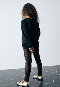 Sudadera negra de corte oversize, combinada con pantalones negros ajustados y zapatos dorados brillantes. El pelo rizado cae por la espalda. Suelo cubierto de confeti.