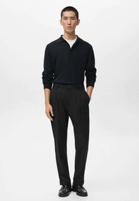 Man staand in een zwart langemouw polo, zwarte op maat gemaakte broek en zwarte schoenen, met één hand in zijn zak tegen een witte achtergrond.