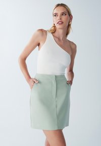 Top bianco a costine con una spalla, abbinato a una mini gonna verde chiaro con tasche laterali e una texture liscia.