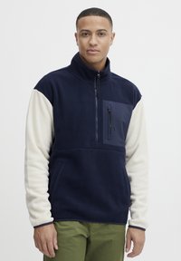 Pull en polaire avec col zippé, corps bleu marine, manches crème et poche poitrine. Présente des textures contrastées et des accents de couture.