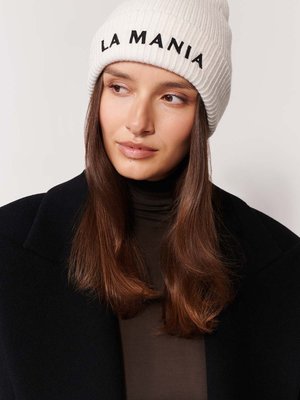 Krémoová rebrová čiapka s čiernym vyšívaním "LA MANIA". Modelka má na sebe tmavý kabát a hnedý top, s dlhými hnedými vlasmi.