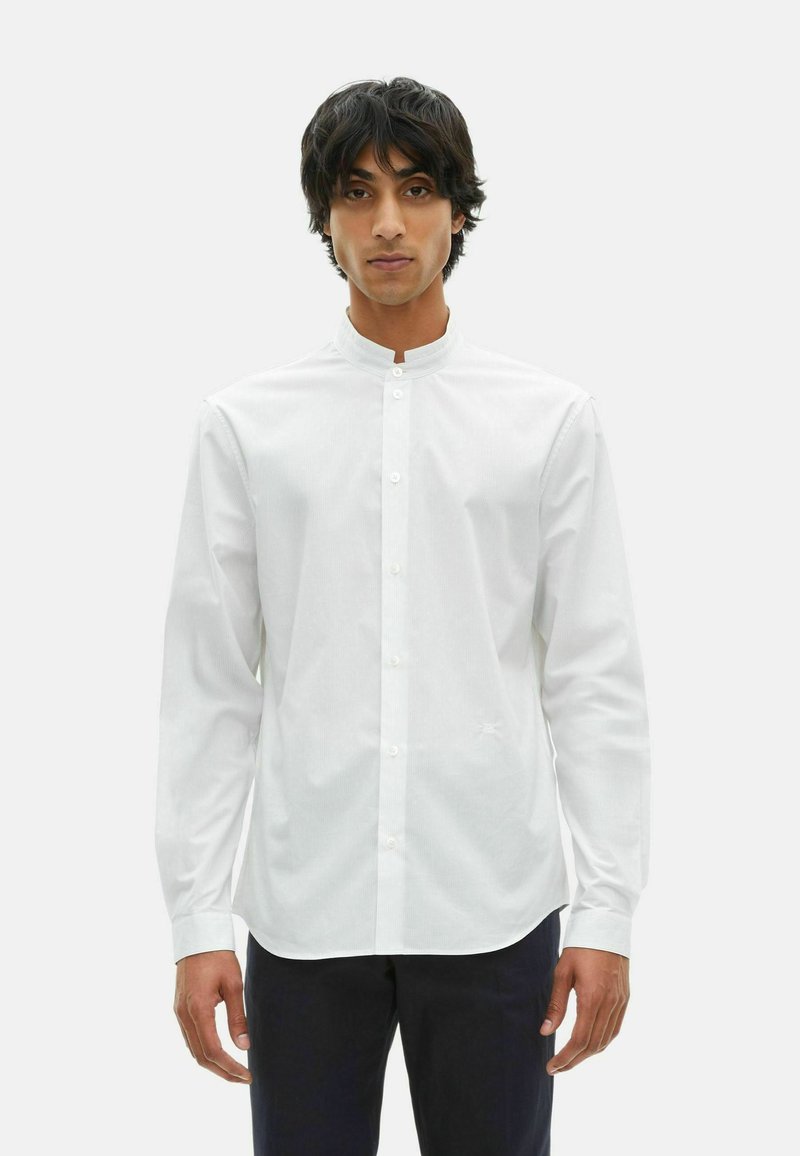 Man met halflang donker haar, gekleed in een witte mandarin kraag knoopshirt en donkere broek, tegen een simpele lichte achtergrond.