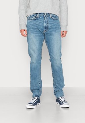 Man draagt lichtblauwe jeans, een grijs shirt met lange mouwen en marineblauwe sneakers met witte veters, staand tegen een effen achtergrond.