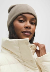 Beige strikket hat og en cremet pufferjakke med høj krave, der har store knapper. Glat tekstur med en afslappet pasform.