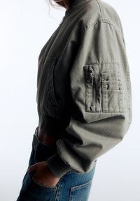 Chaqueta bomber corta de color verde oliva hecha de algodón, con un bolsillo con cremallera en el brazo, puños acanalados y textura suave. Combinada con vaqueros azules.