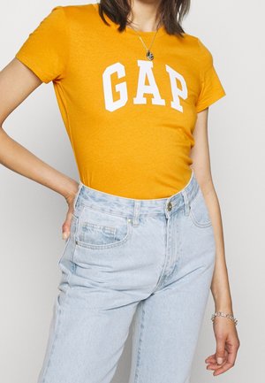 Camiseta de algodón amarilla con el logo "GAP" en blanco, combinada con unos pantalones vaqueros de tiro alto de mezclilla azul claro que tienen un cierre frontal de botones y bolsillos laterales.