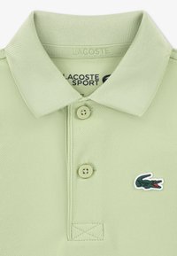 Ανοιχτό πράσινο πόλο μπλουζάκι με ριμπ γιακά, τρία μπεζ κουμπιά και μικρό λογότυπο κροκόδειλου Lacoste στο αριστερό στήθος. Υφή υφάσματος.