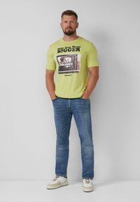 Limoengroene t-shirt met een grafische print van een oude televisie en tekst, gecombineerd met blauwe denim jeans en witte sneakers met beige accenten.