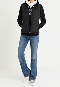 Schwarze gesteppten Puffer-Weste über einem schwarzen Hoodie, kombiniert mit blauen Schlagjeans und grauen Sneakern. Lässiges, geschichtetes Outfit mit lockerer Passform.