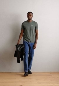 Tricou verde oliv cu logo, blugi albaștri și jachetă din piele neagră. Modelul poartă încălțări negre, mergând pe un podea din lemn, pe fond neutru.
