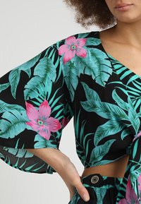 Femme portant un crop top noir avec des feuilles tropicales vertes et des fleurs roses, associé à un pantalon taille haute assorti, main posée sur la hanche.