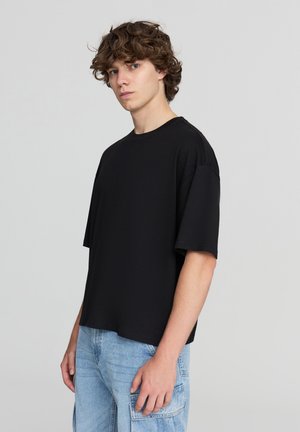 T-shirt in cotone nero con vestibilità rilassata, maniche corte, scollatura rotonda e design delle spalle senza cuciture. Abbinato a pantaloni cargo azzurri chiari.