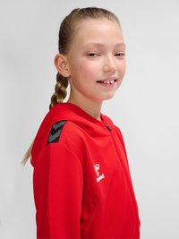 Młoda osoba o blond splecionych włosach ubrana w czerwoną bluzę z kapturem i zamkiem marki hummel, lekko uśmiechająca się, sfotografowana na jednolitym jasnym tle.