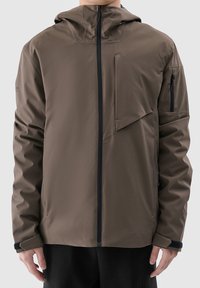 Homme portant une veste à capuche marron avec une fermeture éclair noire et une poche zippée sur la manche, debout bras détendus le long du corps.