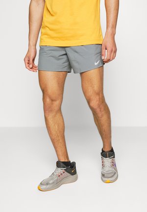 Mann trägt graue Nike-Shorts, gelbes Shirt und graue Laufschuhe mit schwarzen und orangefarbenen Details, steht vor weißem Hintergrund.