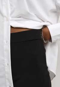 Personne portant une chemise blanche rentrée dans un pantalon noir taille haute, avec une main dans la poche et un bracelet en chaîne argenté au poignet.