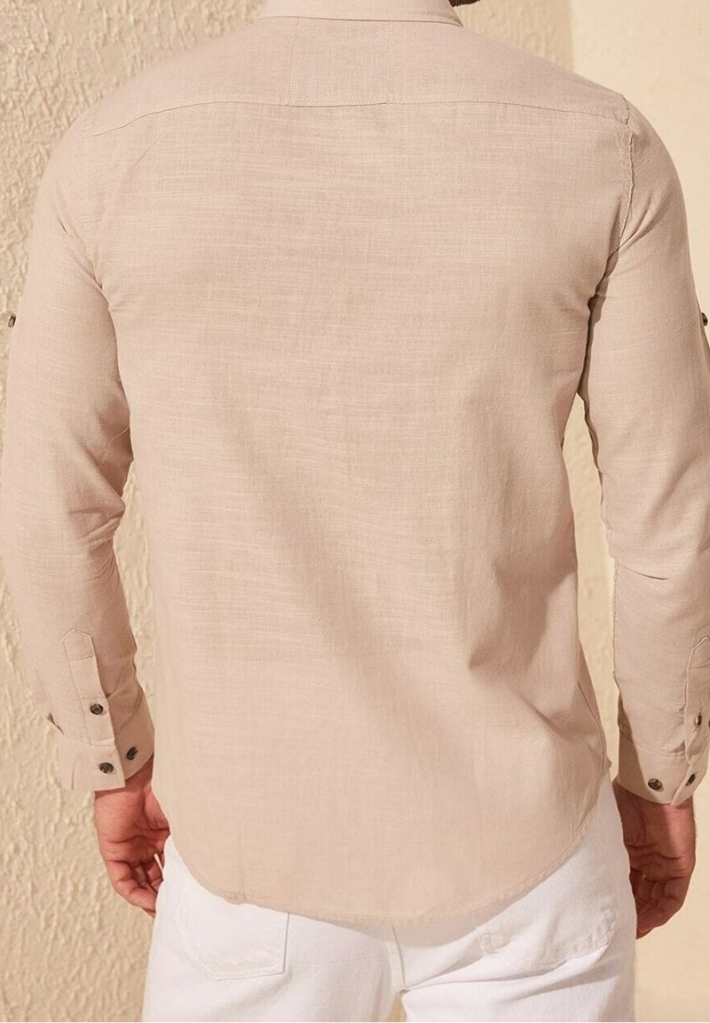 Camisa de manga larga color beige claro hecha de tela texturizada, con puños abotonados y dobladillo curvado. La vista trasera muestra el detalle del yugo en los hombros.