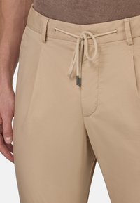 Beige, lahki hlače z vrvico za pas, z gladko teksturo in stranskimi žepi. Okrašene z kovinskimi konicami na vrvici.