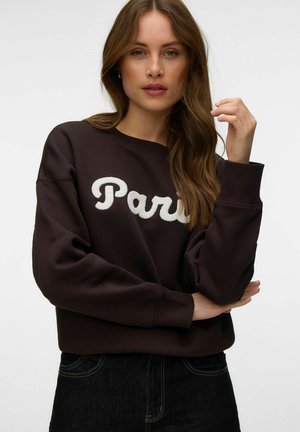 Femme aux longs cheveux bruns portant un sweat-shirt foncé avec "Paris" en lettres blanches, posant avec une main près de son visage sur un fond uni.