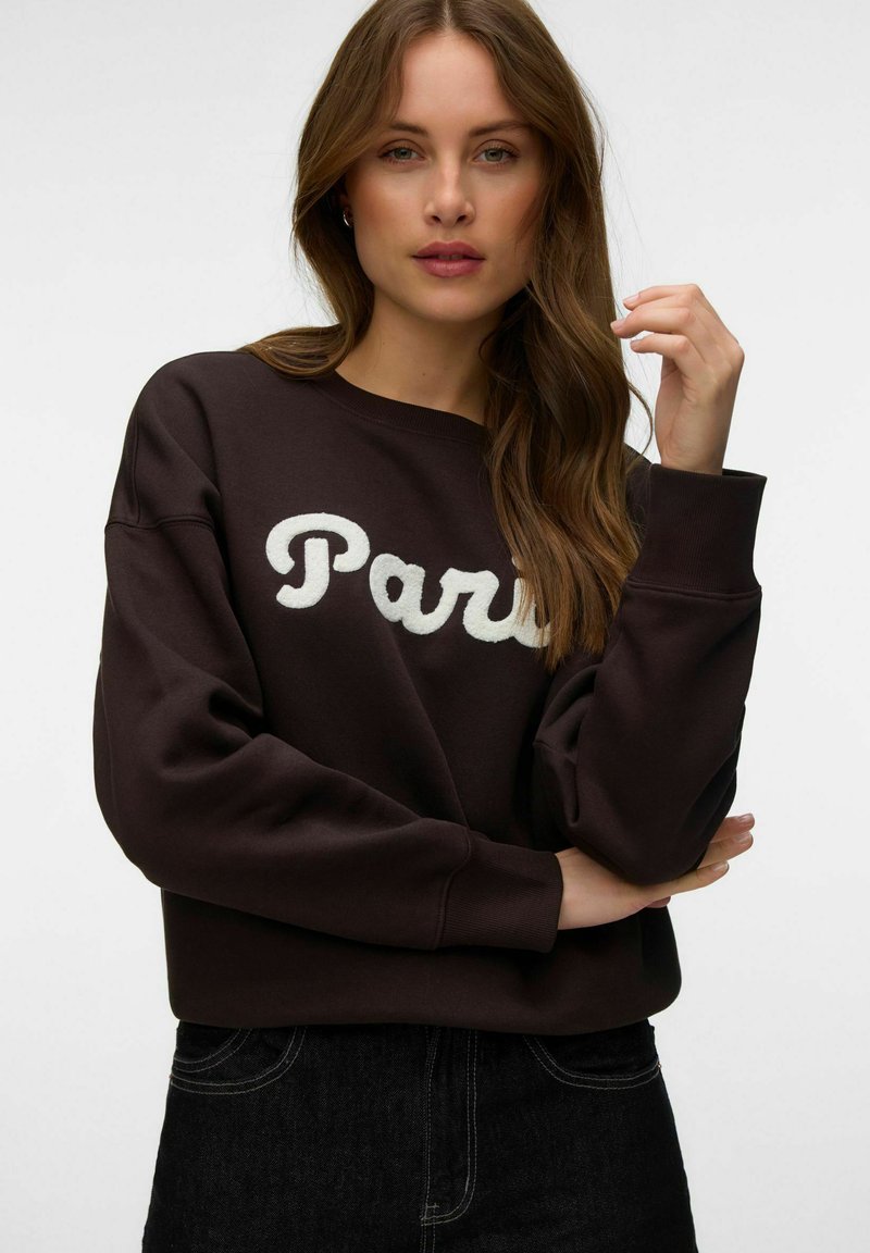 Femme aux longs cheveux bruns portant un sweat-shirt foncé avec "Paris" en lettres blanches, posant avec une main près de son visage sur un fond uni.