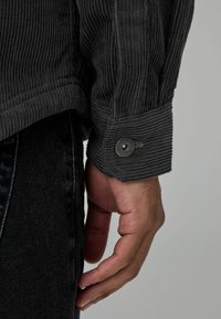 Sort kordfløjljakke med rillet tekstur, med en knap ved cuffen. Hånd hviler mod et par sorte jeans.
