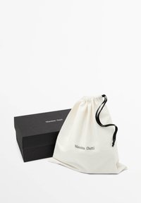 Sac en coton à cordon de serrage couleur blanc cassé avec le logo "Massimo Dutti" imprimé, à côté d'une boîte rectangulaire matte noire, le tout sur un fond blanc.